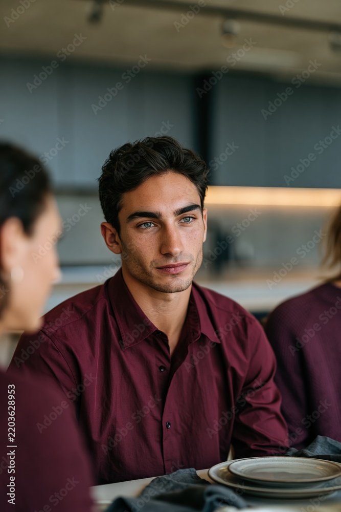 Fototapeta premium Young man facing camera at dining table in burgundy shirt for lifestyle portrait background and social gathering banner