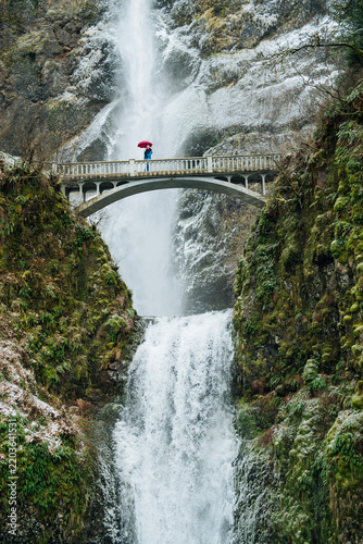 White person with umbrella standing on bridge over multnomah falls