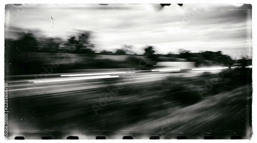 A streaky black and white photo of a road at dusk with car light trails. The image is blurry and gives a sense of speed and movement