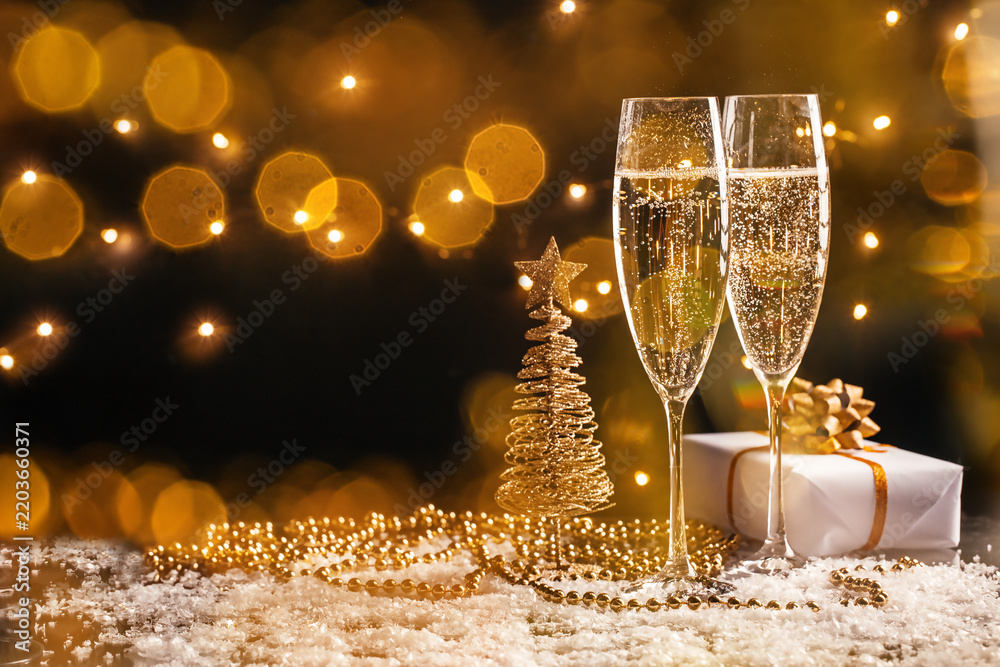 Obraz premium Two champagne flutes beside golden wire Christmas tree and wrapped gift on glittering snowy surface with warm bokeh lights, festive holiday still life arrangement