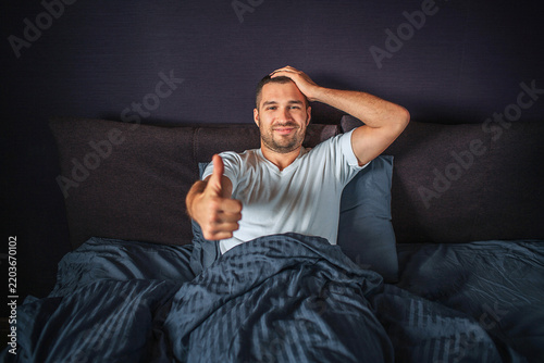 Funny young man sitting on bed and smiling. He shows big thumb up on one hand and hold another one on head. Guy is satisfied and happy.