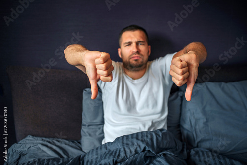 Unhappy and negative young man sits on bed and looks on camera. He holds his big thumbs down. Guy is upset. He is in bedroom.