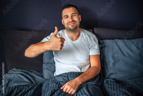 Positive young man sitting on bed and looks on camera. He smiles and shows big thumbs up. Guy is in bedroom.