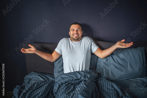Nice and positive young man sitting on bed. He is covered with dark-blue blanket. He keeps hands aside of body. Guy smiles. He is happy.