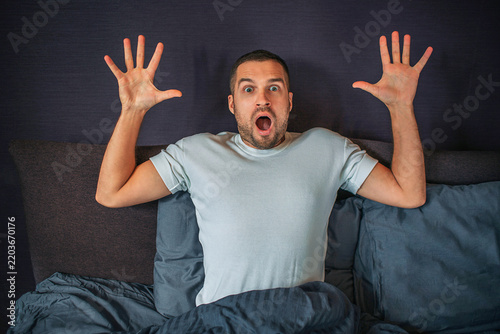 Wondered young man sits on bed and looks on straight on camera. He holds his hands up. Guy is scared and frightened.