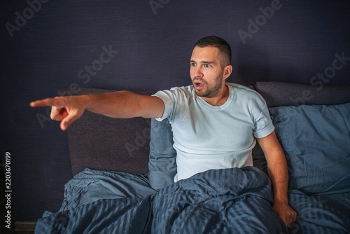 Amazed and wondered young man sitting on bed in bedroom and looks to left. He points with finger. Guy is ecxited.