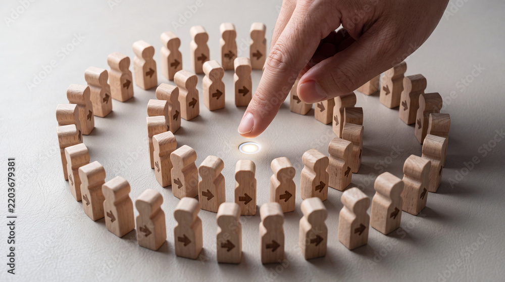 Obraz premium Close up of a hand pointing at a glowing center point surrounded by wooden human figures with arrows pointing inward, representing leadership, team goal, target audience, or human resources concept.