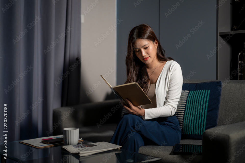 Obraz premium Woman Reading Book on Couch in Modern Living Room with Dark Walls and Sofa
