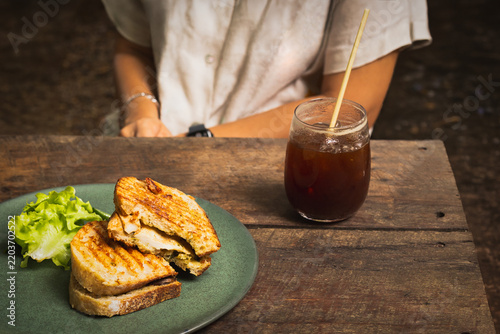 Konstfotografi Woman having grilled chicken panini sandwich with glass of iced Americano on wooden table