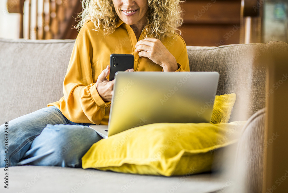 Fototapeta premium Woman on couch using smartphone and laptop to compare home insurance quotes, relaxed on a yellow cushion while browsing policies and handling finances online from home easily.