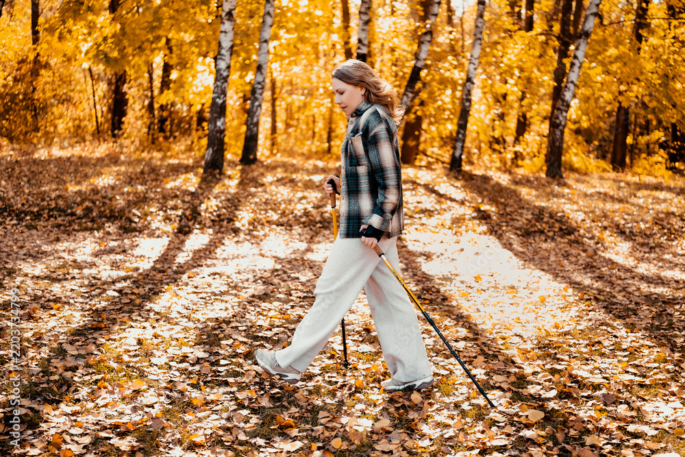 Fototapeta premium Adult woman in autumn woodland with trekking poles hiking outdoor. Fall nature walking for physical and mental health. Finding peaceful relaxation and enjoying solitude outside city.