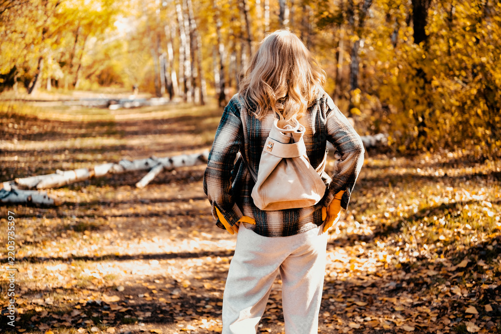 Obraz premium Back view of woman in autumn nature with stylish backpack. Fall outdoor hiking and mental health restoration. Leaving busy city life for peaceful solitude journey through yellow forest.