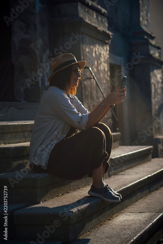 Happy female traveler sitting on historic stone steps, using a smartphone at golden hour. Relaxed city lifestyle, solo travel, summer vacation and urban exploration