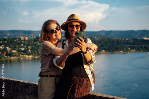 Two smiling female lesbian travelers embracing at a scenic river viewpoint, enjoying friendship and love during summer vacation with panoramic hills and water under a beautiful evening sky