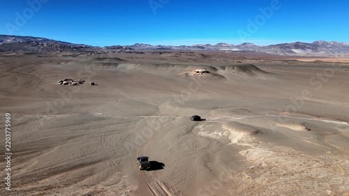Off-road SUVs cross dry pumice plains in Campo de Piedra Pómez with distant Andean mountains under clear blue sky, useful for themes of adventure travel, remoteness and exploration.