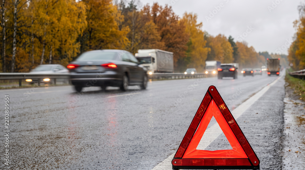 Fototapeta premium Warning triangle on wet highway with cars passing in rainy weather 