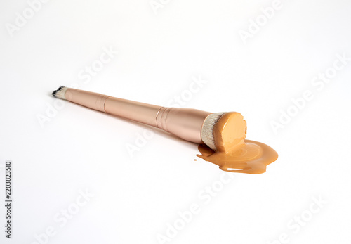 A rose gold makeup brush and liquid foundation that has pooled onto a white surface.