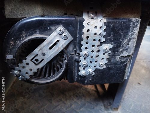 Foto Close-up of an improvised DIY repair on a cracked black plastic blower fan housing, reinforced with perforated metal mounting straps and metal screws to hold the broken parts together