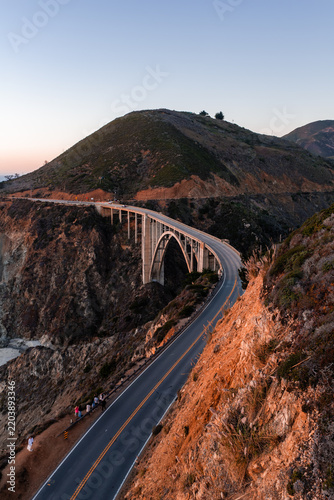 Bixby Creek Bridge curves along Highway 1 on the rugged Pacific coast in Big Sur, California, with distant tourists standing by the roadside at sunset as warm light illuminates the cliffside.