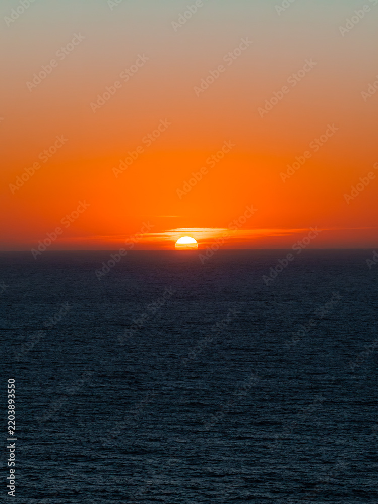 Obraz premium Sun sets directly into the Pacific Ocean horizon viewed from the Big Sur coast in California, illuminating the evening sky with warm orange tones above gentle waves.