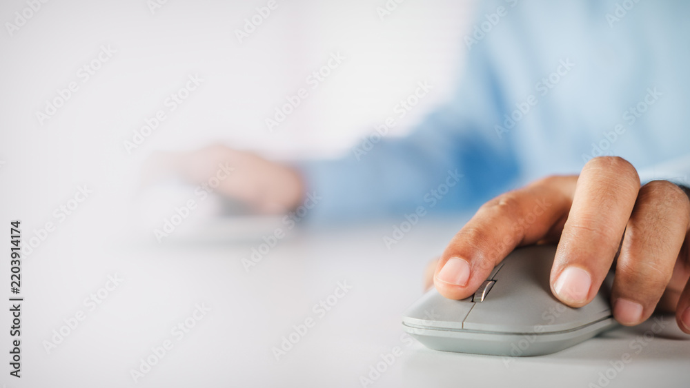 Fototapeta premium Hand of a businessman holding a grey wireless mouse on a white desk with copy space, concept of working with computer, digital technology, online business, and professional productivity.