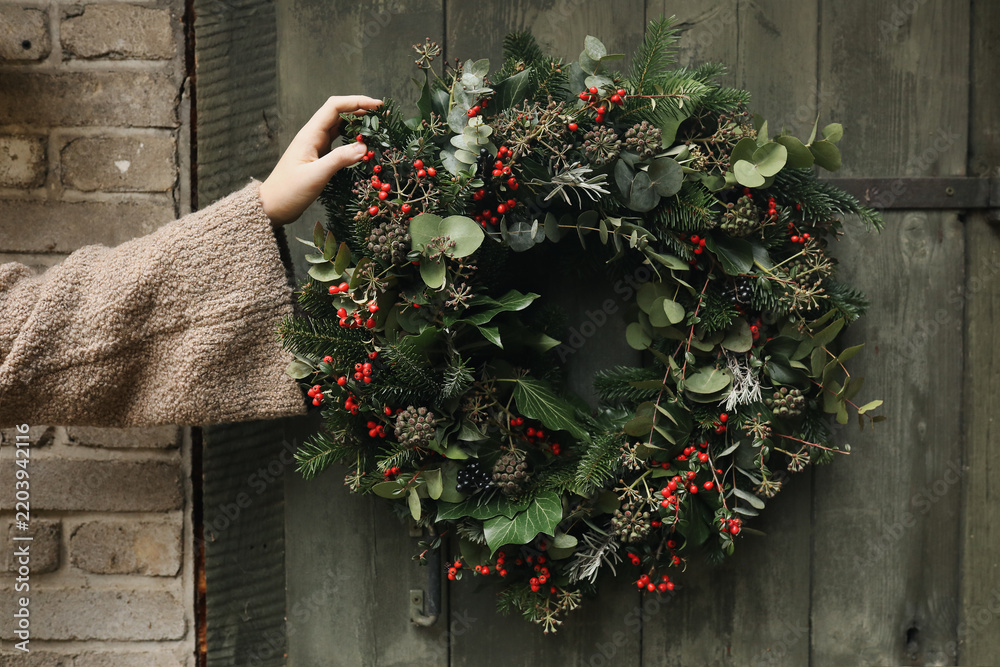 Obraz premium Female Hand in sweater Holding Handmade Christmas Wreath with Evergreen Foliage, Red Berries, Ivy, Eucalyptus on Blurred Old Wooden Door. Festive Winter Holiday Decoration, Natural floral home decor