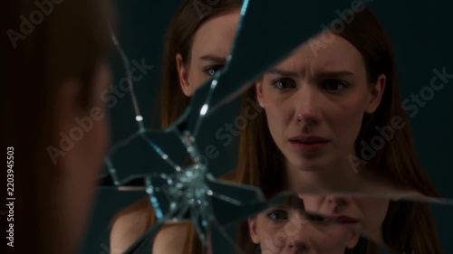 Young woman stares into a shattered mirror. Her expression shows shock and dismay. Glass fragments reflect her face clearly. The dark background contrasts her features