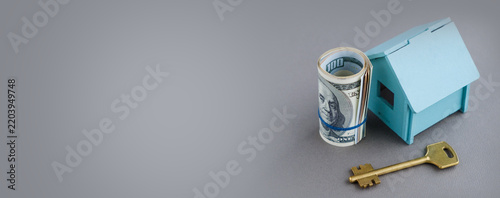 Real estate and mortgage photography featuring a small blue house model, a roll of US dollar banknotes, and a house key on a grey background.