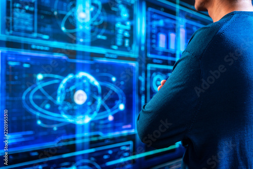 Back view of a man watching glowing blue digital screens with atom symbols, charts and data, showing science, technology and research.