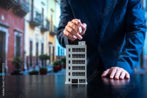 Business professional placing a small office building model on a table, symbolizing real estate investment, urban development, planning and corporate decision-making against a blurred city street.