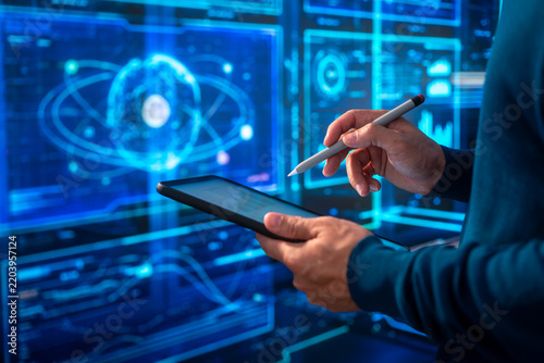 Scientist using a tablet and stylus in front of glowing digital screens with an atom graphic, showing data analysis, research, and modern technology.