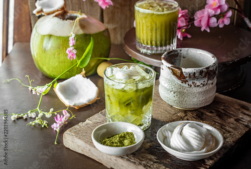 Iced coconut matcha green tea drink served with fresh coconut meat, powder, and bamboo whisk on dark wooden table. Refreshing Asian fusion beverage for summer refreshment menu in cafe.