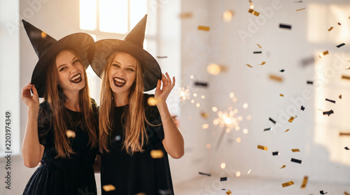two happy young women in black witch halloween costumes having fun at party with firecrackers and confetti on white background funny faces cinematic golden hour lighting professional portrait with cop