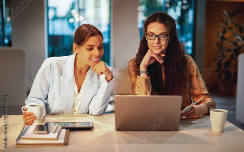 A blonde woman in a light blazer holds a coffee cup, her curly haired colleague in glasses holds a printout, and both smile at a laptop, chins resting on hands.