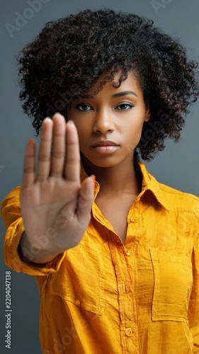 Strong woman confidently raises her hand signaling a pause, embodying empowerment and self-assurance in a stylish yellow shirt against a neutral background.