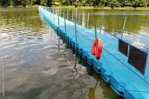 Long blue floating plastic pontoon bridge with a pedestrian walkway and orange lifebuoy stretching across a calm green lake towards a summer forest.