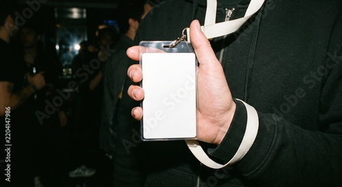 Young man holding a blank vertical plastic event badge with a white lanyard at a night event. Realistic mockup for pass and credentials branding.