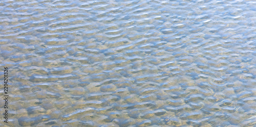 Natural background of clear shallow water flowing over smooth pebbles, with gentle ripples, sunlight reflections and soft blue tones creating a calm abstract texture