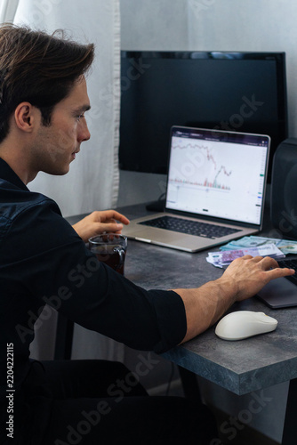 Young man in black shirt analyzes financial data on laptop screen while sitting at desk with cash and a cup of coffee in a modern home office setting