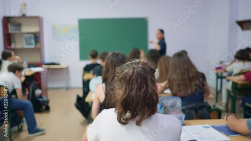 A group of young students sits at desks in a classroom while a teacher stands at the front and a pupil raises their hand