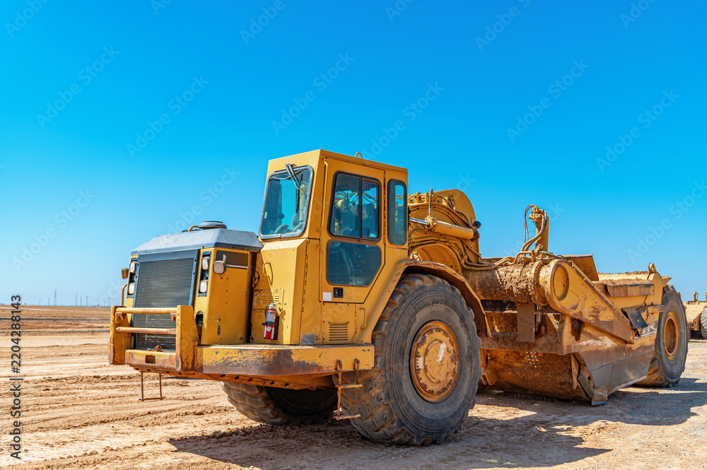 Fototapeta premium Yellow scraper stands on construction site under blue sky. Heavy construction machinery.