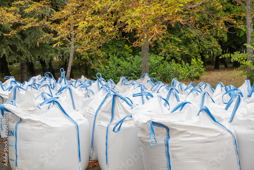 A large arrangement of filled white industrial bulk bags with blue lifting straps sits outdoors beneath early autumn trees on a park construction site