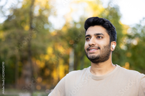 Smiling young man wearing wireless earbuds enjoying audio content while relaxing in a park, representing lifestyle, technology, and connection to nature