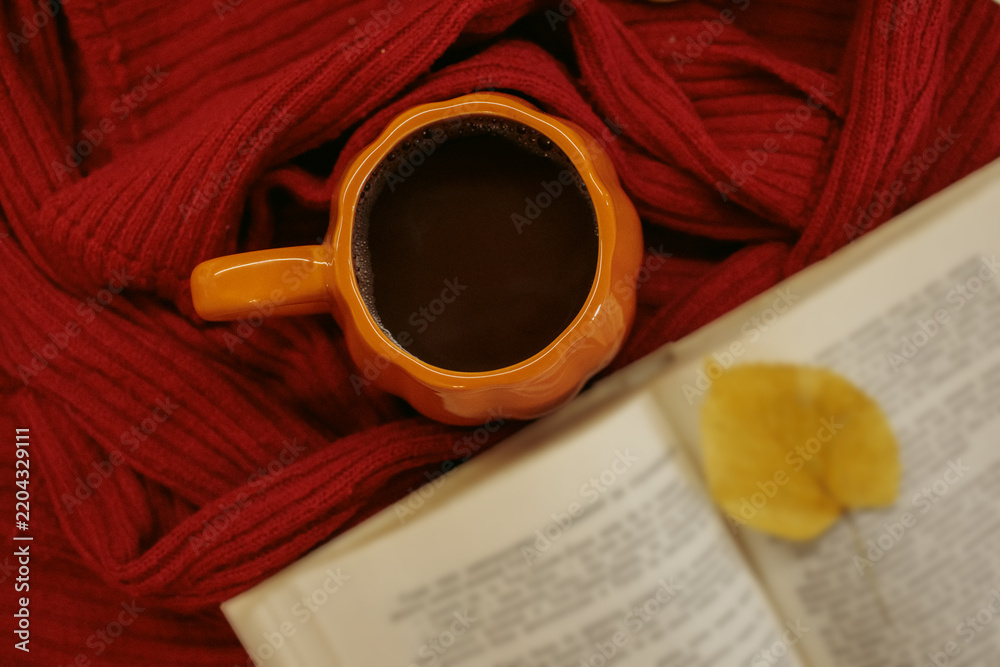 Fototapeta premium An orange mug of coffee resting on a red knitted sweater beside an open book and a yellow autumn leaf, with soft film grain, high quality photo