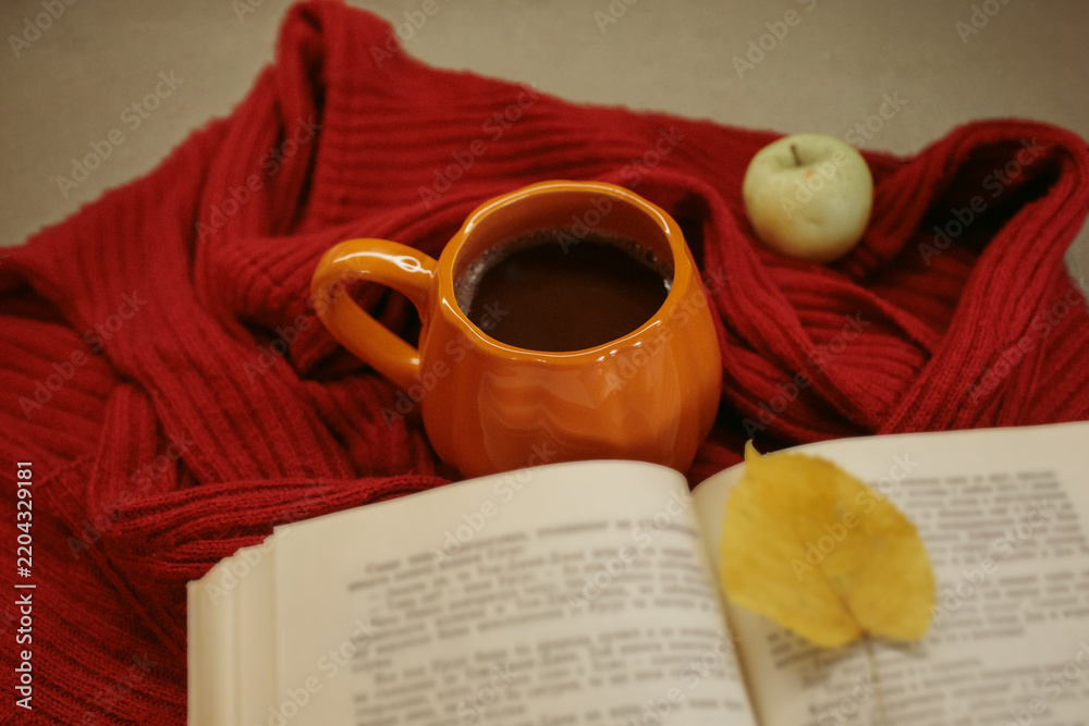Obraz premium An orange mug of coffee resting on a red knitted sweater beside an open book and a yellow autumn leaf, with soft film grain, high quality photo