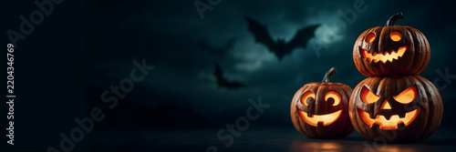 A spooky halloween composition with three carved, glowing jack-o-lanterns stacked and placed on a dark surface against a cloudy night sky with bats