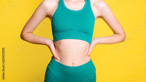 Close-up of slim fit woman waist and torso in green sportswear standing against yellow background