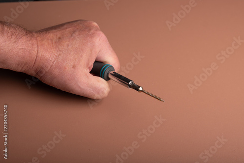 A human fist is holding a screwdriver and a screw is magnetic connectet to the phillips screwhead. A wooden board as background with space for text or digits.
