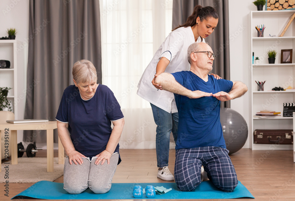 Obraz premium Physiotherapist adjusts the arm and shoulder posture of a senior man during a kneeling rehabilitation exercise while a senior woman rests beside him at home, supporting functional fitness.