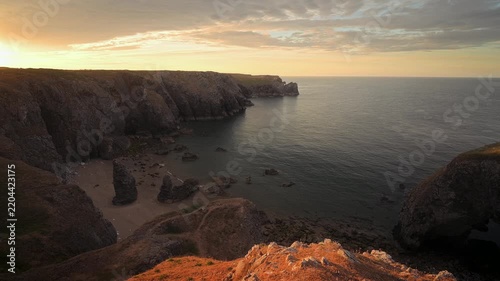 A glowing evening horizon and orange foreground rocks frame the rugged Pembrokeshire coastline and tranquil sea.